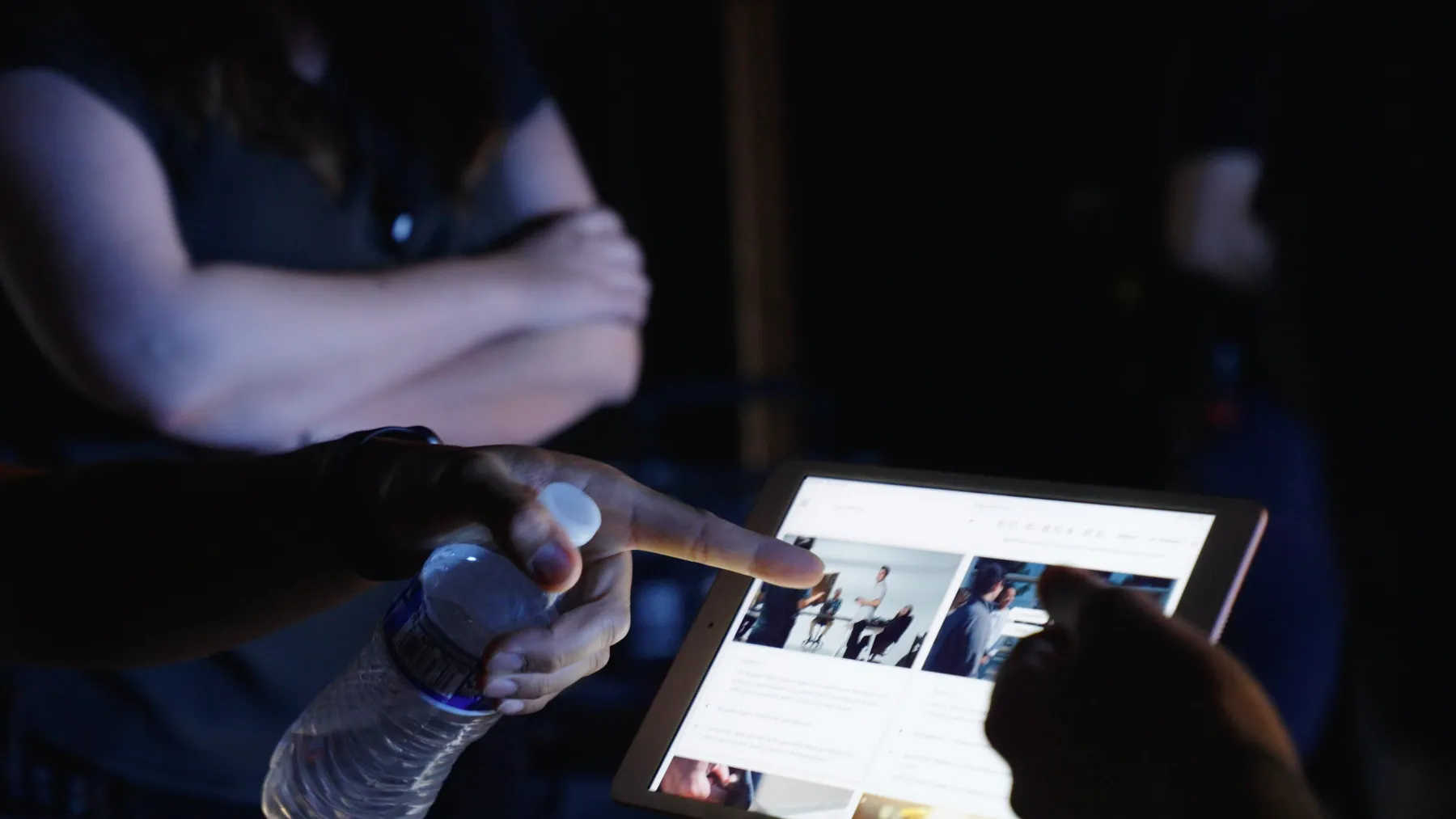 In the dark on set, a hand points at a tablet showing frames from the shoot while someone beside it holds a water bottle.